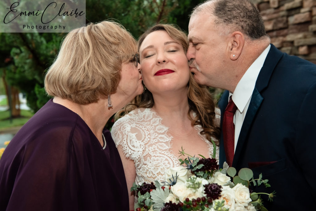 Bride smiles as her parents kiss the sides of her cheeks on her wedding day while making time for family portraits after her ceremony