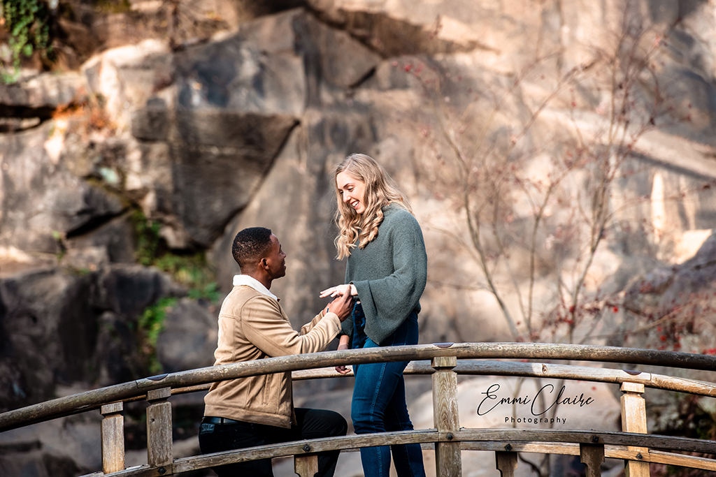 Man in a tan jacket and dark jeans gets down on one knee to propose to his girlfriend outdoors during their winter engagement photoshoot