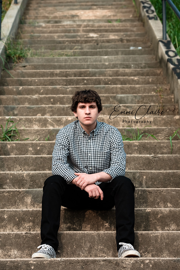 young man in a white and black checkered shirt and black pants sits on a staircase and poses for his senior photos