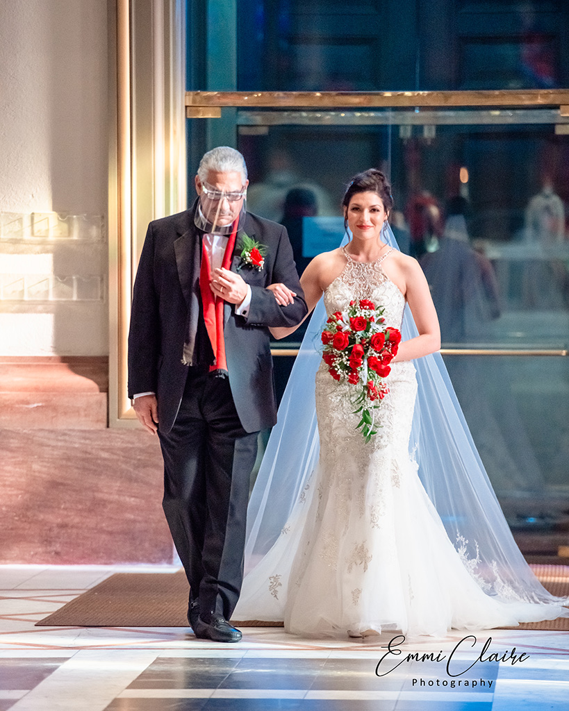 bride in stunning mermaid gown walks down the aisle with her father carrying a bouquet of red roses