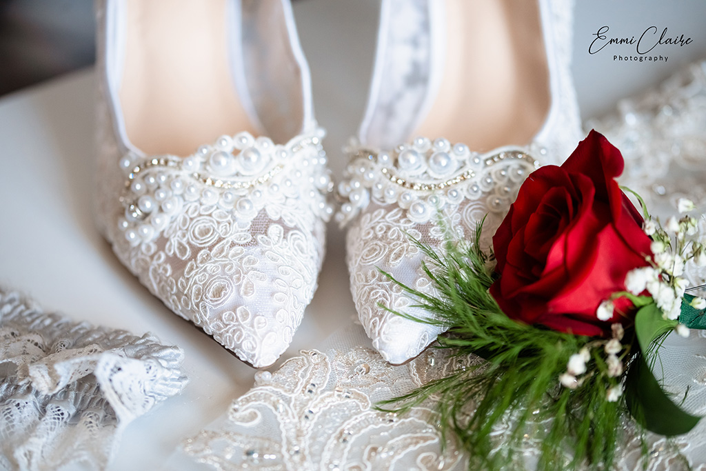 Wedding close up shot of the bride's lace and pearl detailed shoes next to a red rose boutonnière 