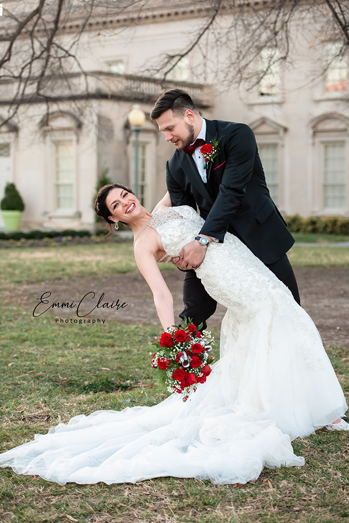 Groom dips his bride while she looks at the camera with a wide smile holding her bouquet of red roses below her in one hand