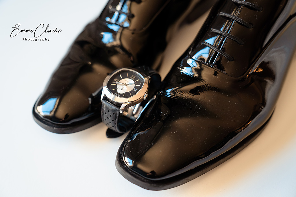close up photo of a groom's shiny, black wedding day shoes with his gold and black watch facing up between the soles of the shoes