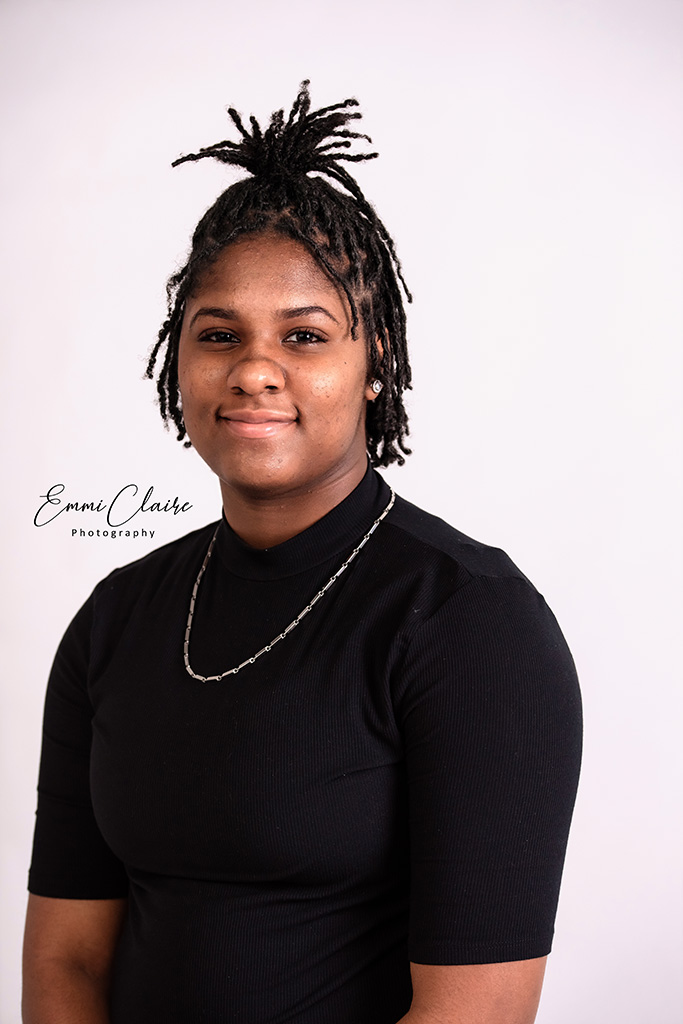 young woman in a black shirt wearing a chain and with her hair half up poses indoors for her senior portraits 