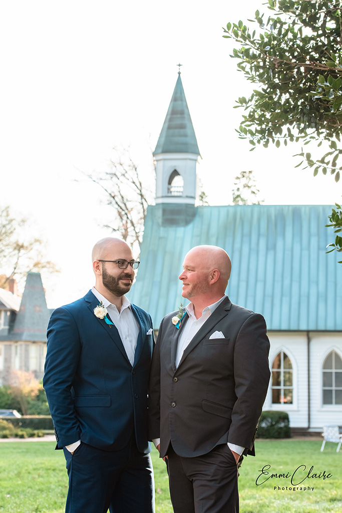 Newlywed couple looks at one another in the eyes and poses in front of a church outside together in their suits 