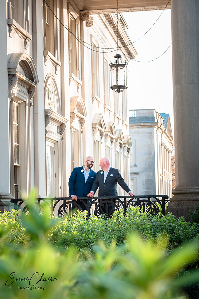 two well-dressed men stand side-by-side holding hands in a beautiful outdoor space with shrubs and greenery in front of them
