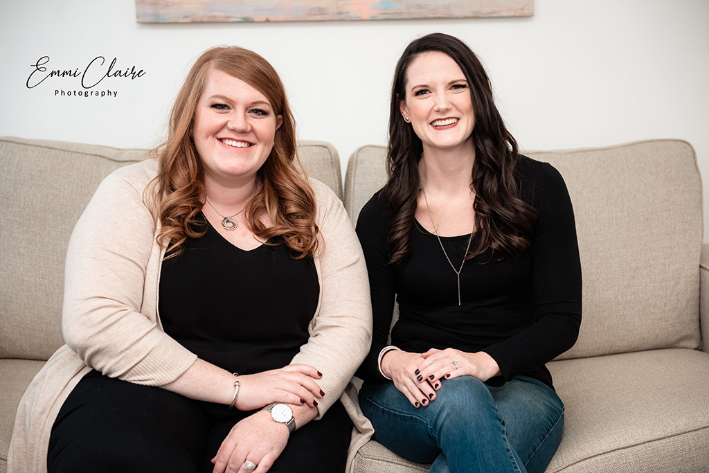 Two therapists sit side by side on a neutral-colored sofa smiling with their hands on their laps 
