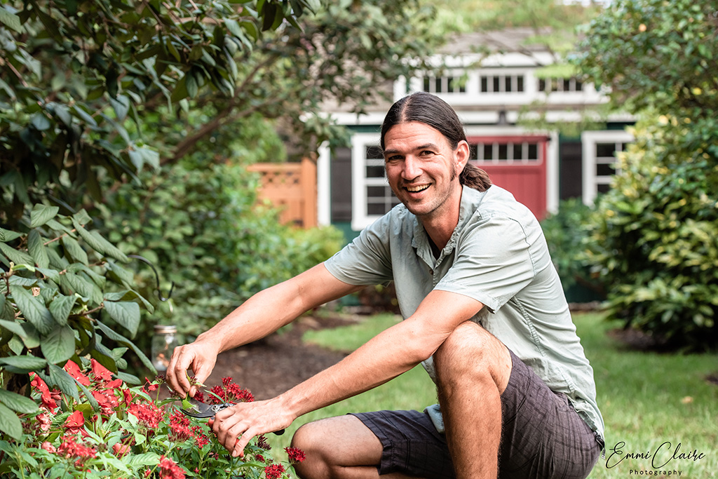 man with his hair in a ponytail kneels down trimming flowers in his garden in front of his home during a personal photoshoot session