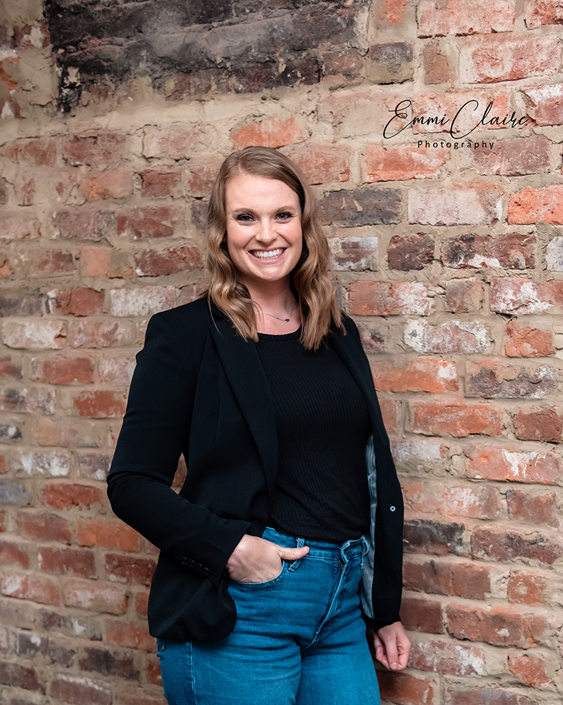 woman poses in a black shirt, black blazer, and jeans in front of a brick wall for photos that will support her small business