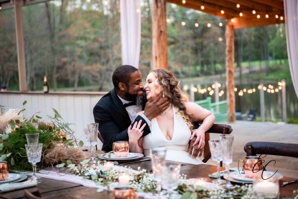 bride and groom sit together at a table during their beautiful outdoor reception with their faces close about to share a kiss