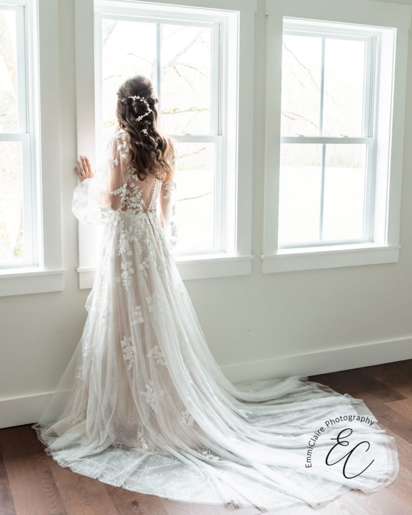 bride standing in a sheer and lace long-sleeved bohemian wedding dress in front of a white window staring outside during a styled shoot