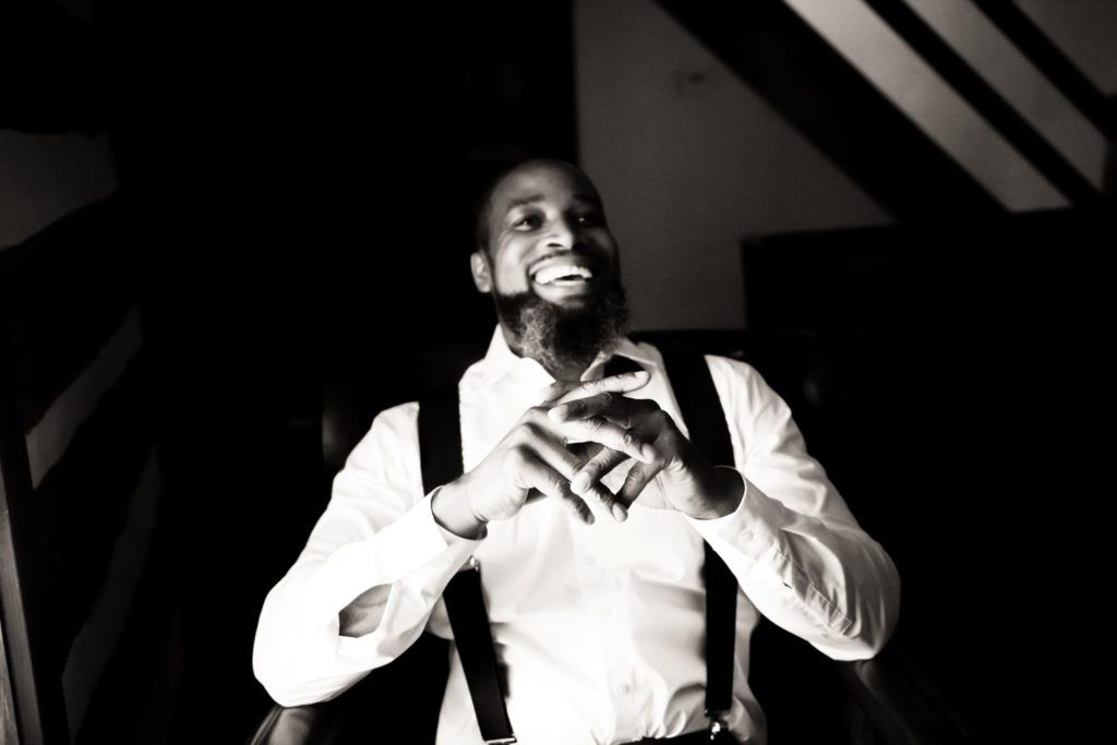 black and white photo of a groom smiling with his hands clasped sitting in his white dress shirt and suspenders