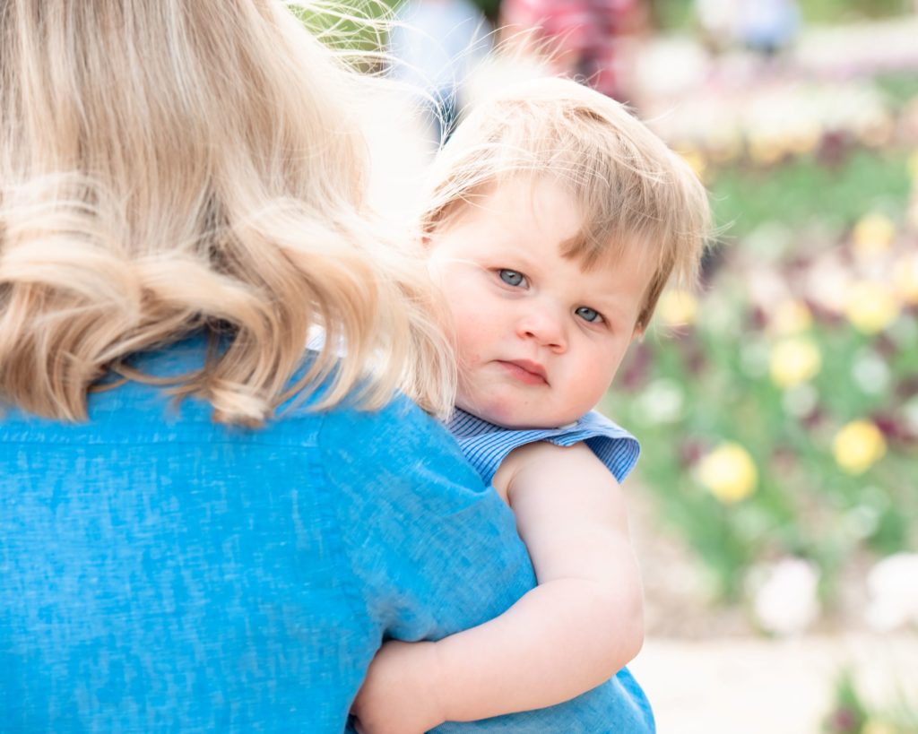 small child dressed in blue stares into the camera and holds onto his mom who has her back faced to the camera as they stand during springtime outdoors for their mini session