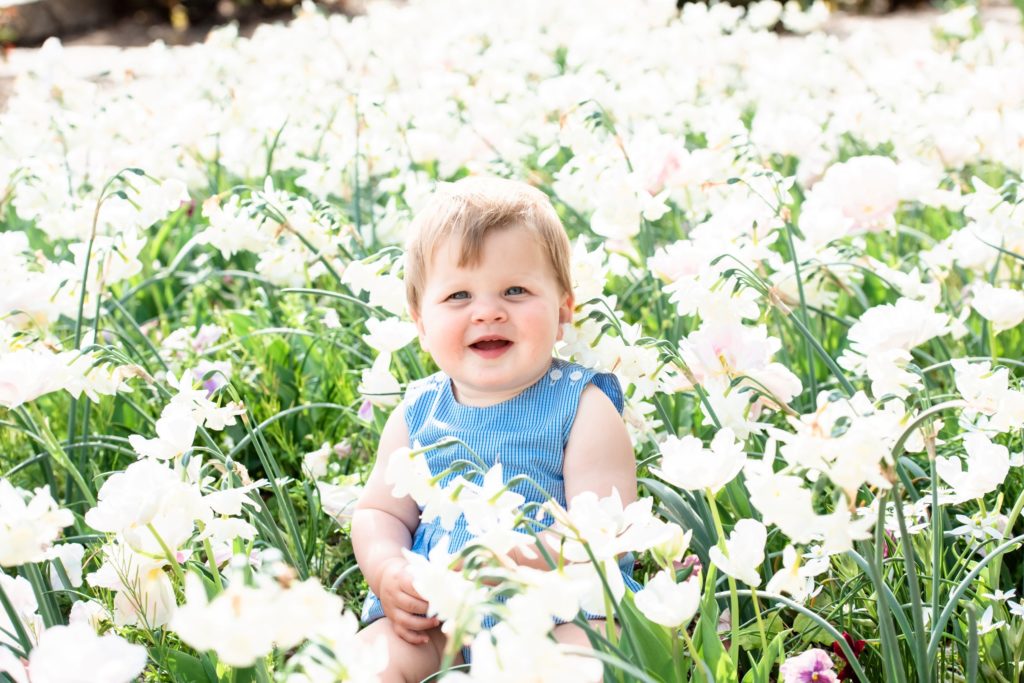 Baby in a cute blue outfit smiles while sitting in the grass surrounded by beautiful white flowers which will make the perfect gift as a photo print for family members
