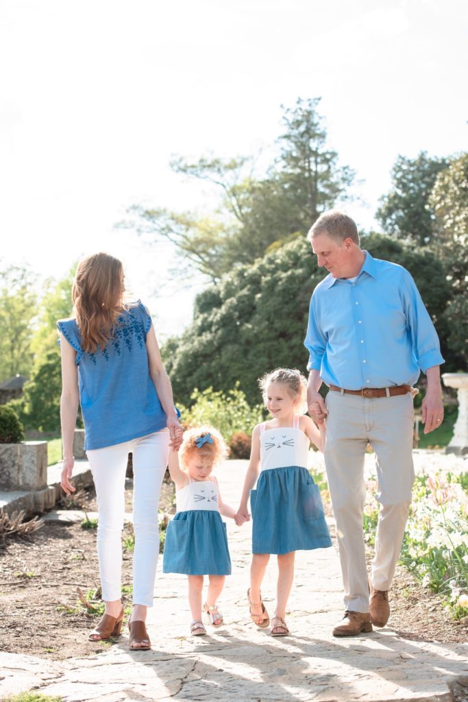 Mom, two young daughters, and Dad all walk holding hands on a beautiful garden path as the two young girls show off their matching blue and white dresses