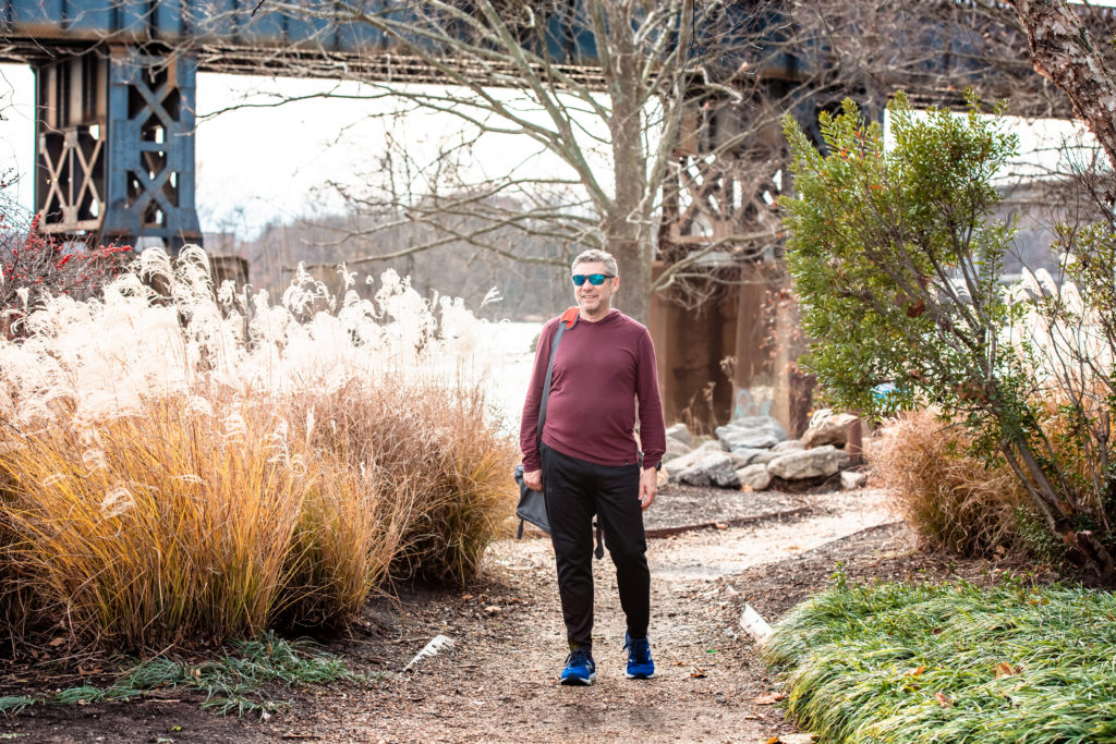 middle-aged man in burgundy hoodie and black sweat pants walks through a park with his sunglasses on during a relaxed photoshoot