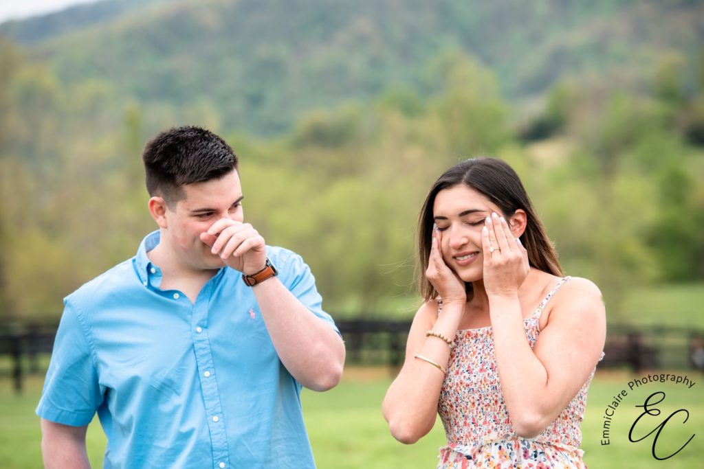 women in a floral jumper wipes away her happy tears after surprised by a proposal from her partner outdoors while her now fiance smiles beside her