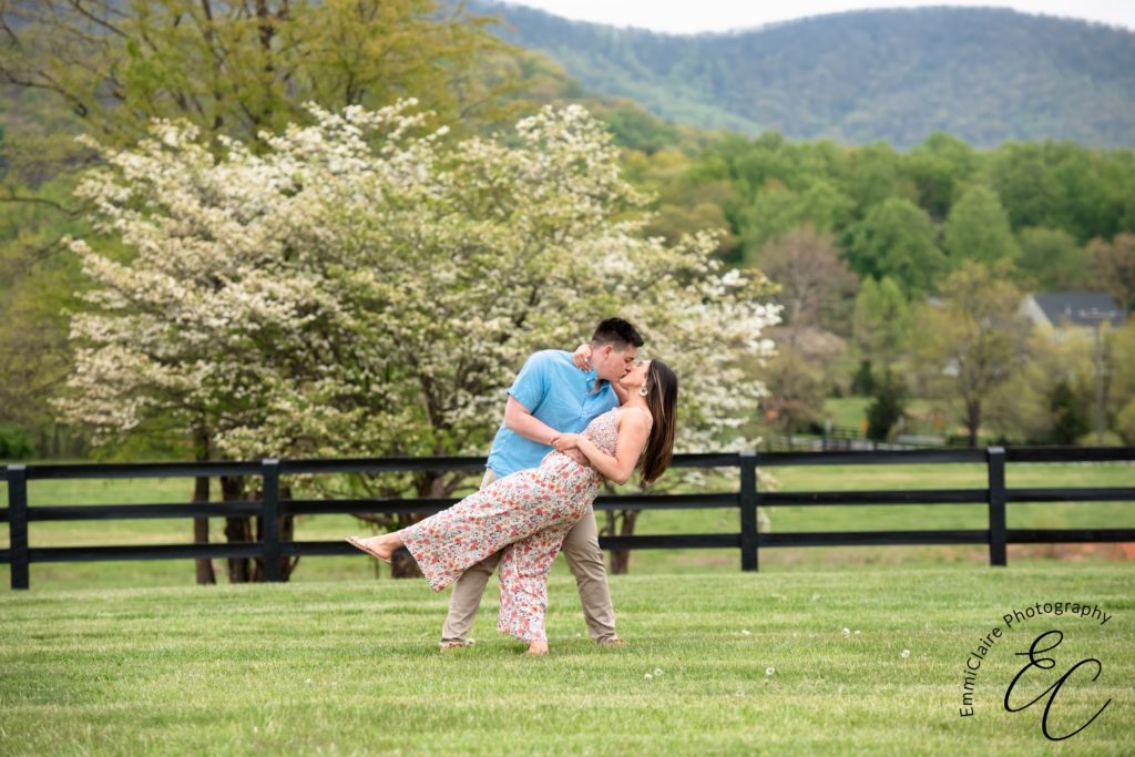 Man lovingly dips his newly-engaged fiance as they stand in the grass together while sharing a kiss during their surprise proposal photoshoot