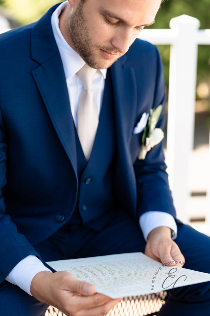 groom sitting outside in his navy blue tuxedo reading a letter from his soon-to-be bride before their ceremony begins