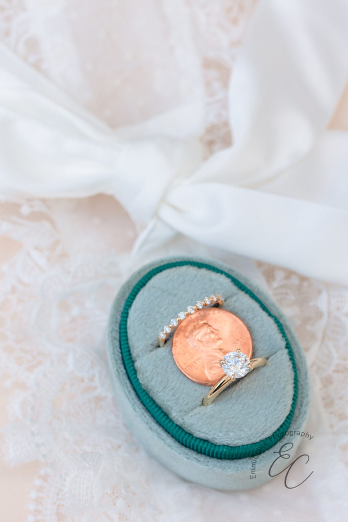 wedding detail shot of a pastel green ring box with the engagement and wedding ring inside separated by a flat laying lucky penny