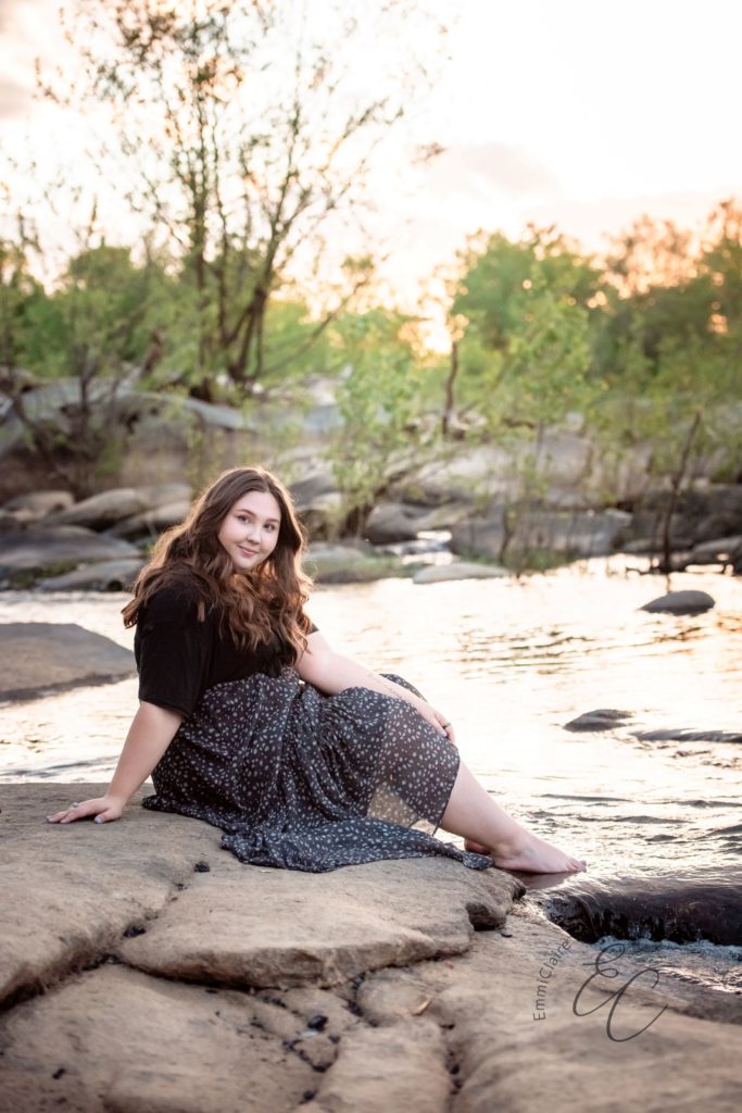 young woman smiles at the camera while sitting on a rock at sunset during her senior portrait session at Belle Isle