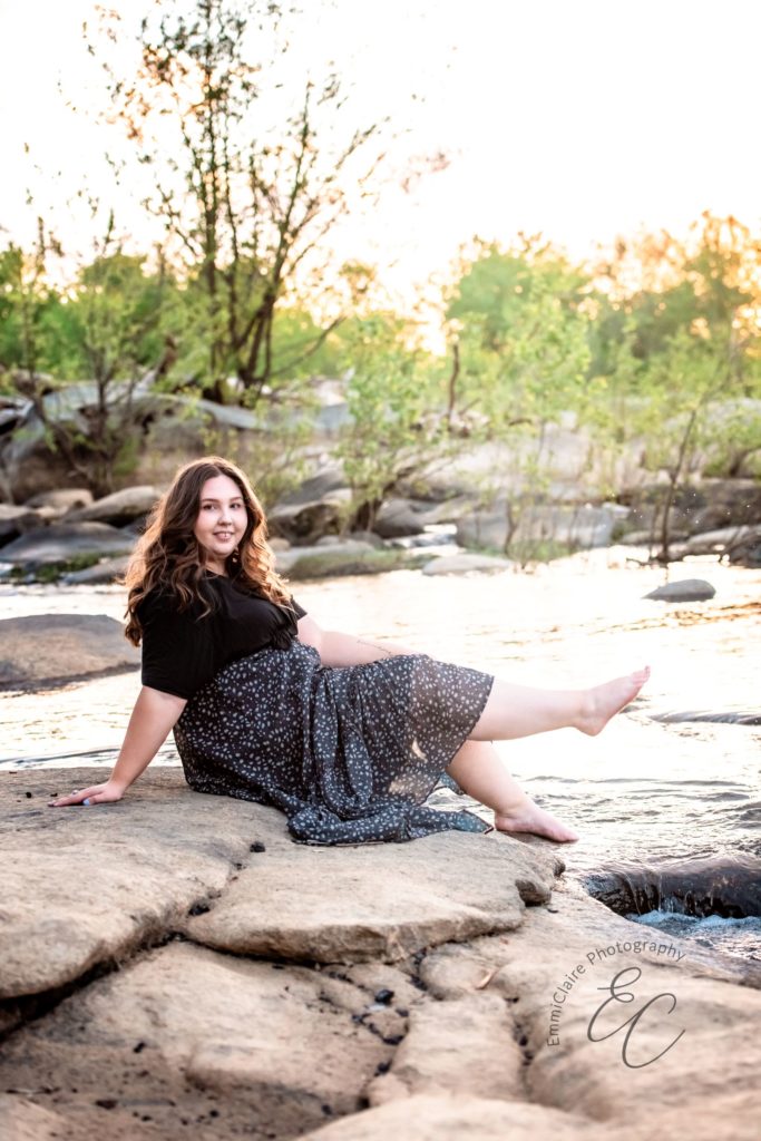 Senior graduate in a black shirt and navy blue skirt poses on rocks with her bare feet dipping in the stream next to it