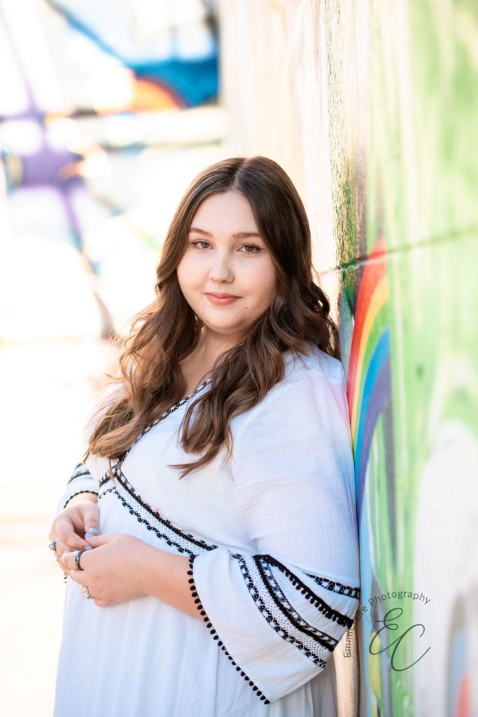 Young woman poses in a white dress next to a colorful wall during her senior portrait session 