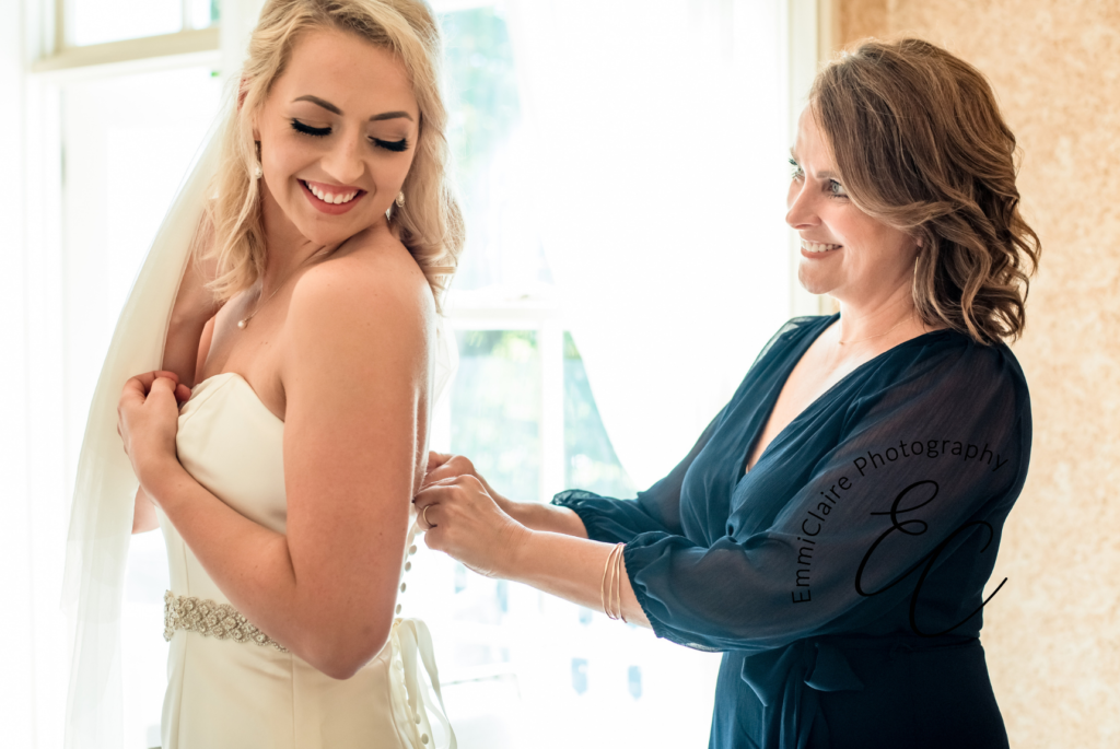 Mother in a navy blue dress standing behind her daughter helping her button up the back of her wedding gown while the bride is smiling and looking back