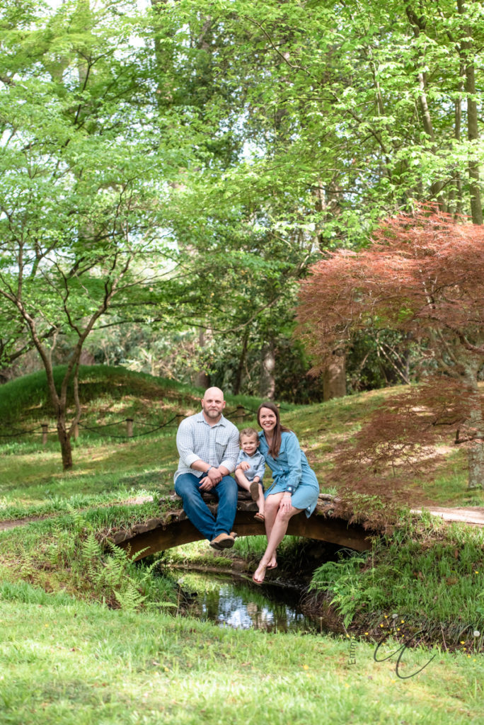 A young boy sits between his father and mother with their feet hanging over a small bridge that spans over a creek during their family photoshoot