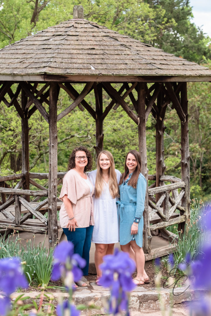 A mom and her adult daughters standing in front of an outdoor gazebo with arms around one another and smiling for a portrait together