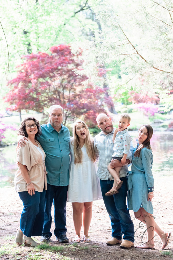 Six family members stand together in front of a pond making silly faces at the camera during their outdoor family photo session 