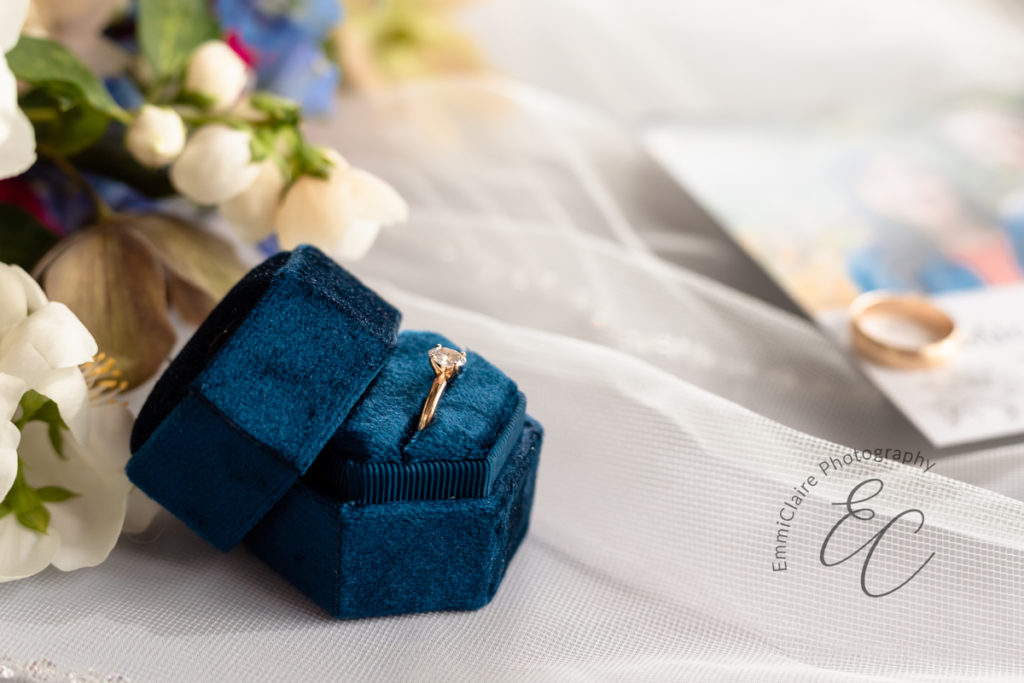 detail shot of a gold wedding ring in velvet navy blue box surrounded by other small details of the cottage wedding in the background