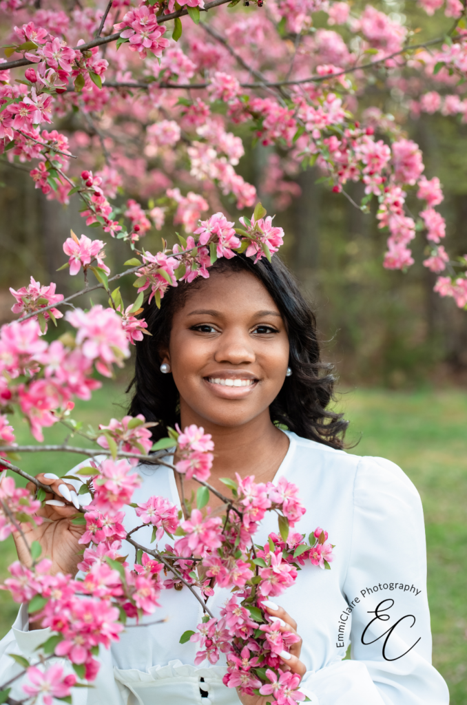 young woman smiles in pearl earrings and a white dress next to blooming cherry blossom tree flowers during her senior portrait session