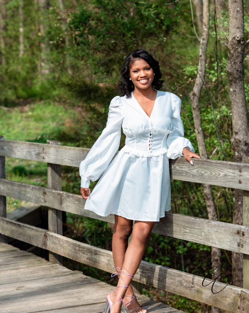 young woman standing outside on a wooden bridge smiling in the sun wearing a white dress during her senior photoshoot in Virginia