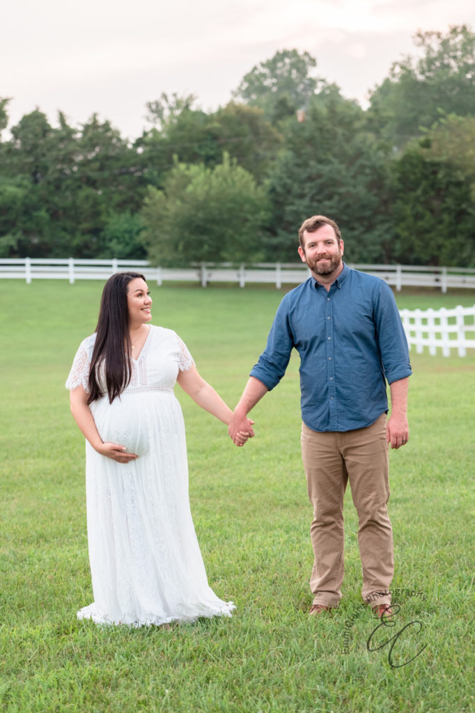 expectant mother in a long, white dress holds hands with her husband in a beautiful green space during their outdoor maternity photoshoot