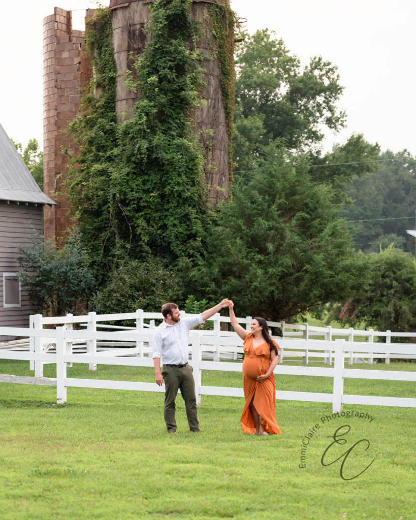 pregnant woman in a long, orange dress dances with the father-to-be in the grass during their maternity photoshoot