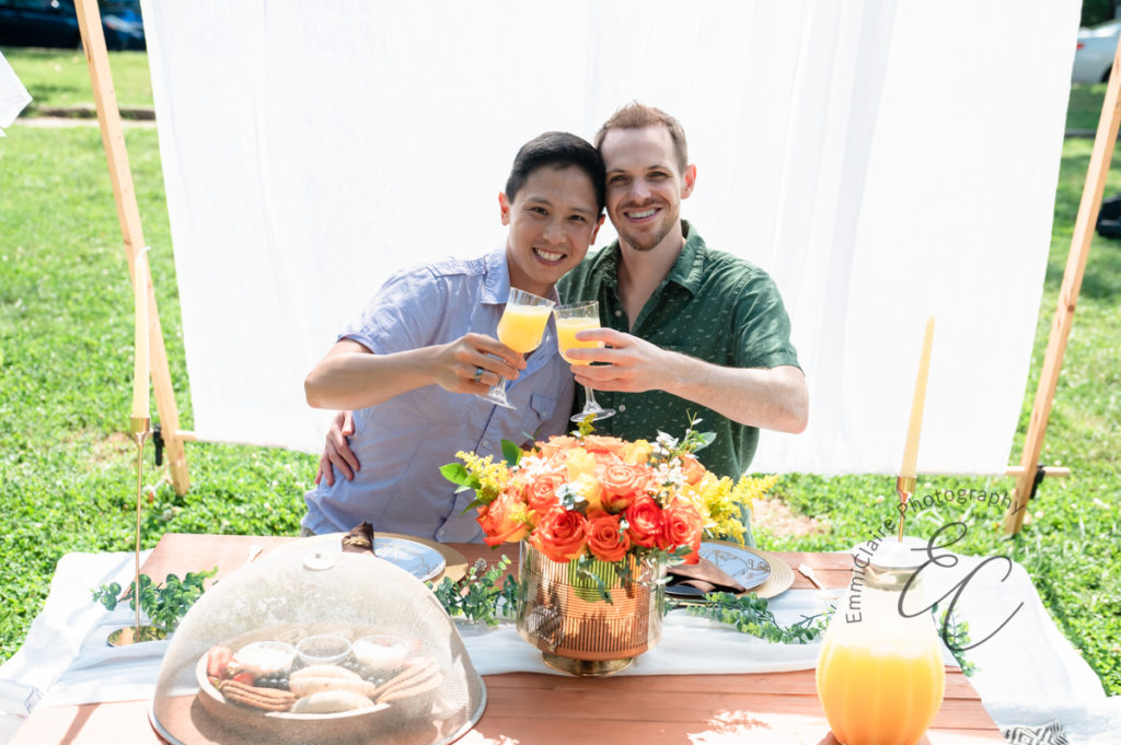 newly engaged couple does a cheers with orange juice during their picnic engagement photoshoot