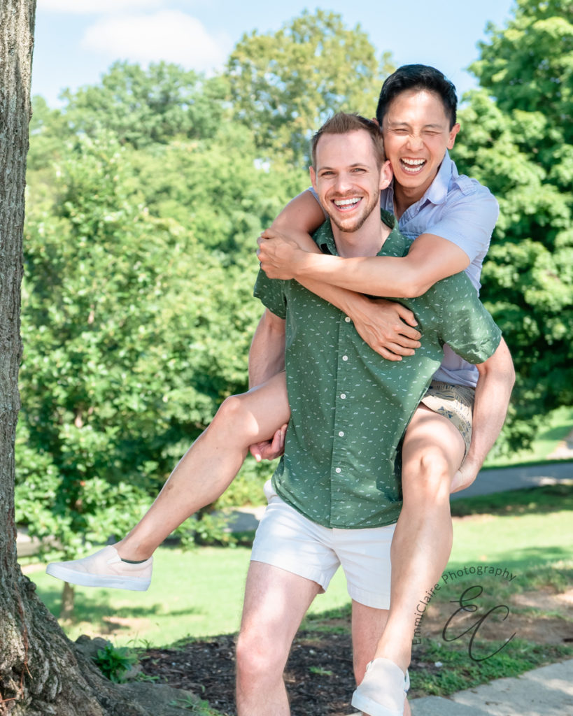 man in a green shirt smiles while giving his partner a piggyback ride in a beautiful park on a sunny day after they just got engaged