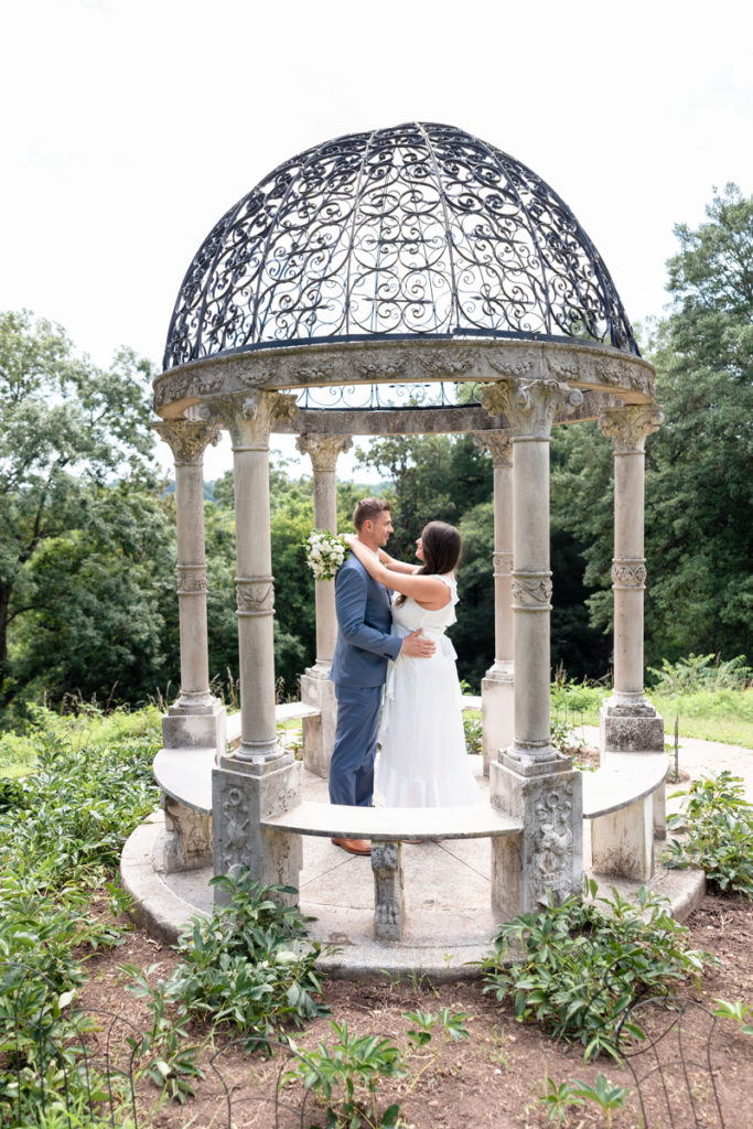 newlywed couple stands together underneath a beautiful outdoor marble gazebo sharing an embrace