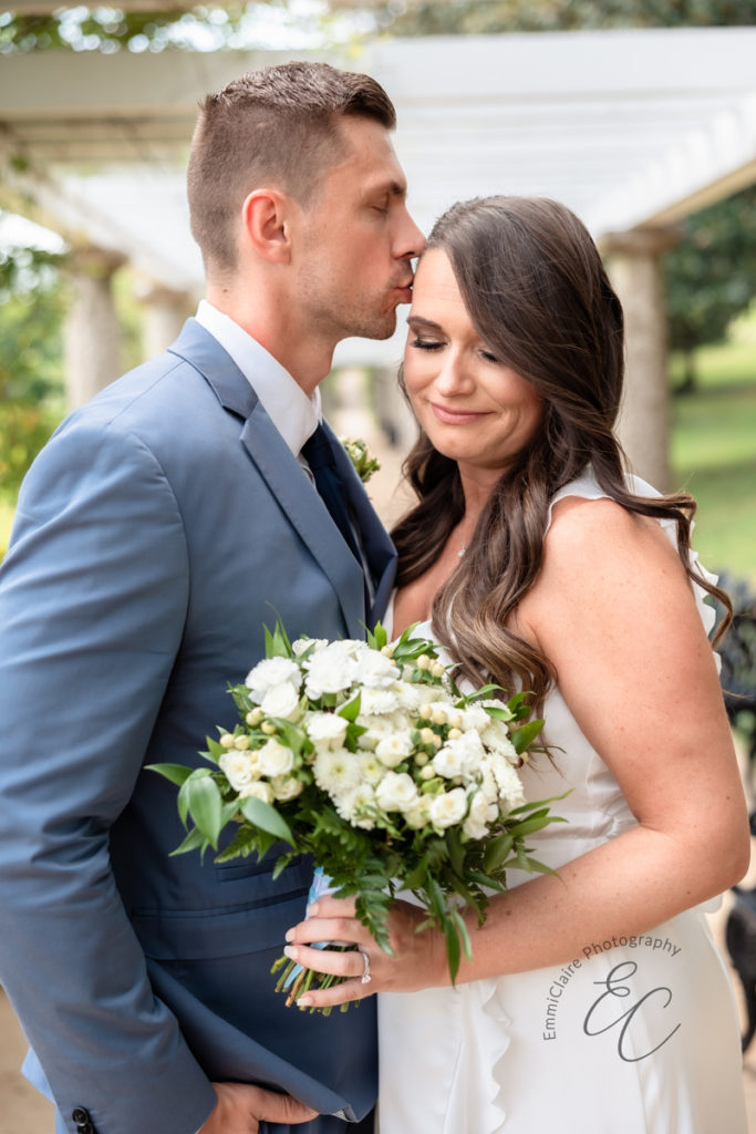groom kisses his bride on the forehead while she holds her white bouquet during their quaint courthouse wedding