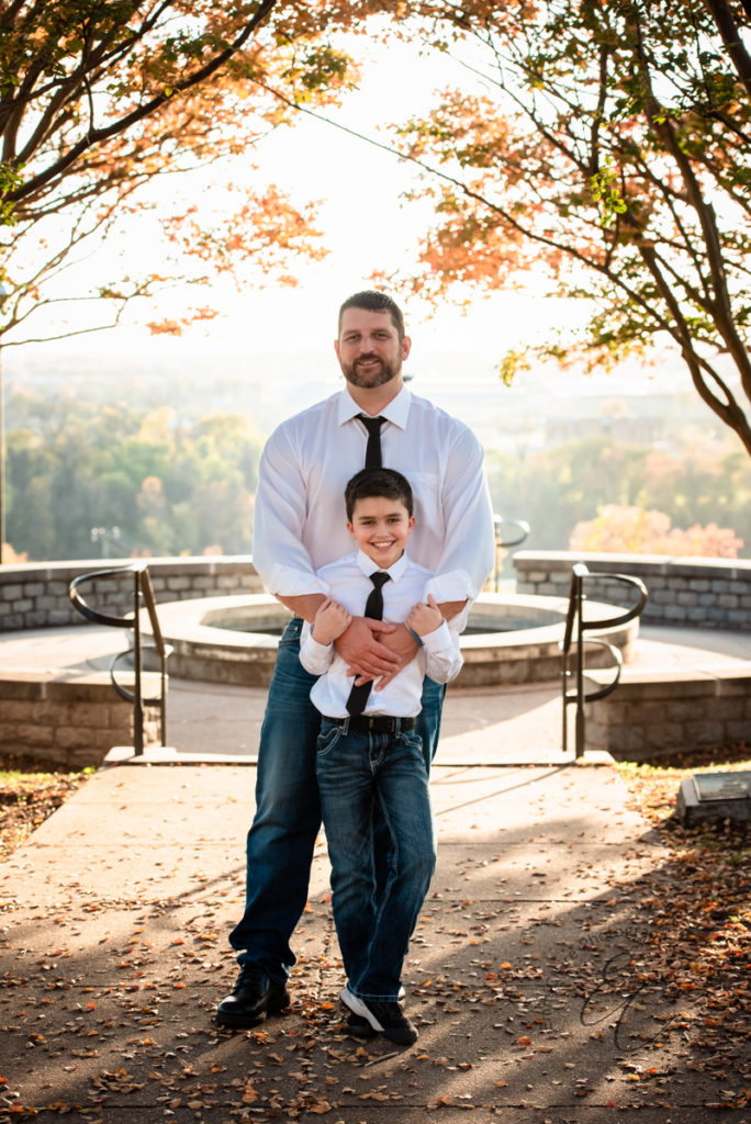 father and son in matching jeans, white dress shirt, and black ties stand together outside in the fall for a photo together