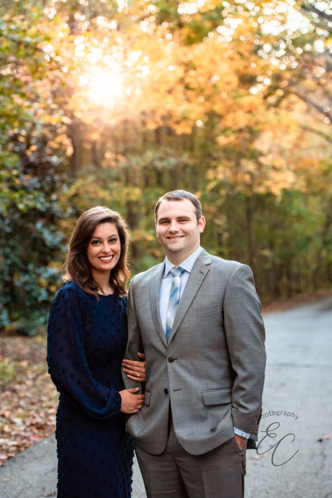 well-dressed couple stands together arm-in-arm on an outdoor path smiling during their fall mini photoshoot session