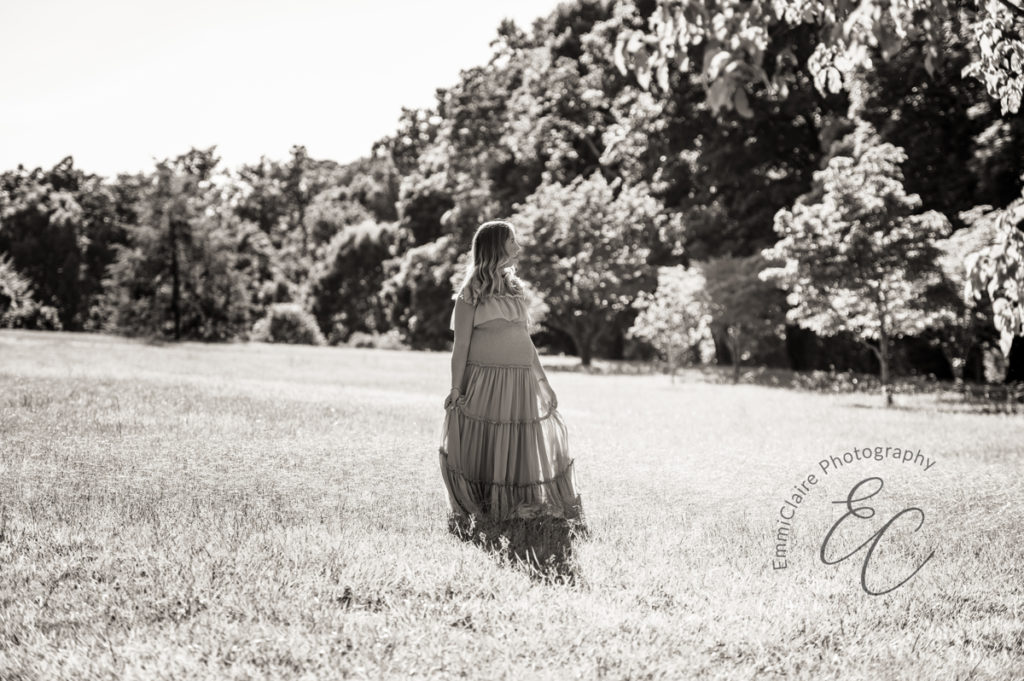 black and white photo of a pregnant woman in a long, flowing dress walking through a beautiful field surrounded by trees