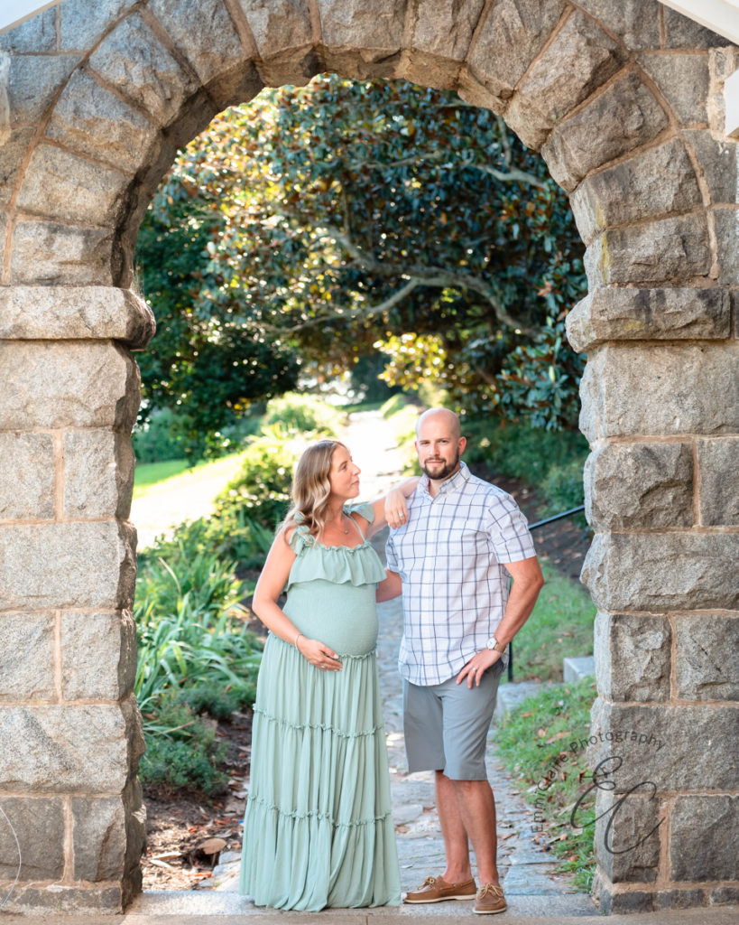 Expectant couple stands under a beautiful outdoor stone archway to capture her pregnancy through beautiful maternity photos