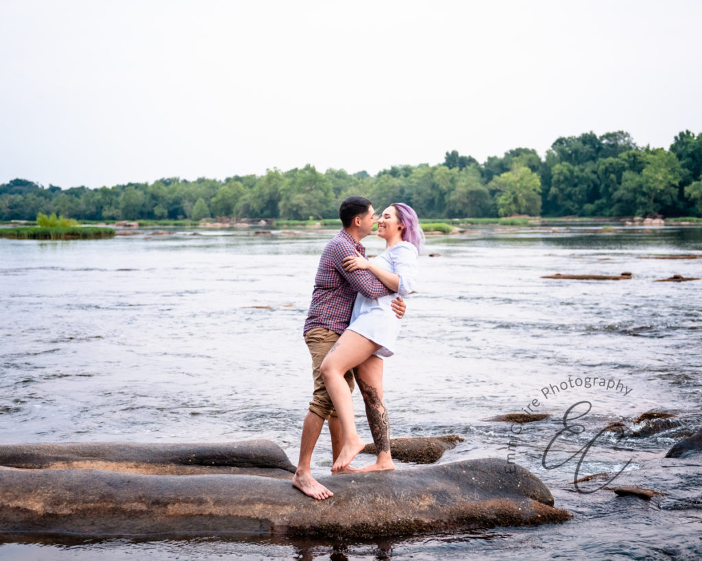 couple laughs and smiles while standing on a rock in bare feet beside a flowing river during their adventure engagement photoshoot