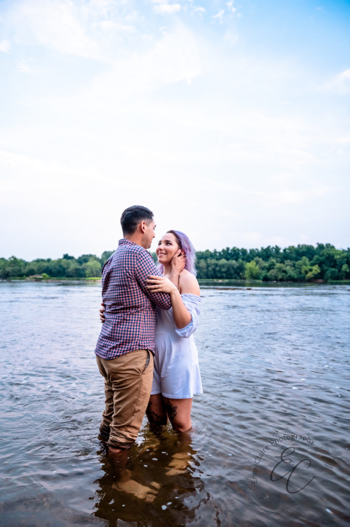 woman with purple hair in a white jumper stands in knee-deep water with her fiance as they look at one another smiling
