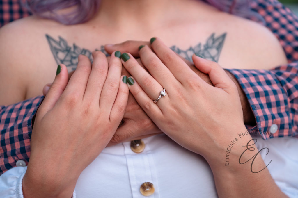 close up shot of a mans arms wrapped around his fiance while she places her hands over his and shows off her engagement ring