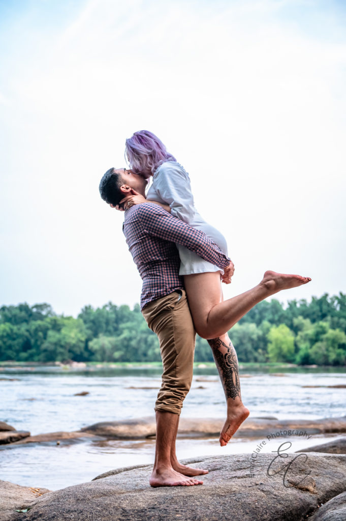 woman jumps into her fiances arms and wraps her arms around his neck as they kiss in bare feet beside a flowing river