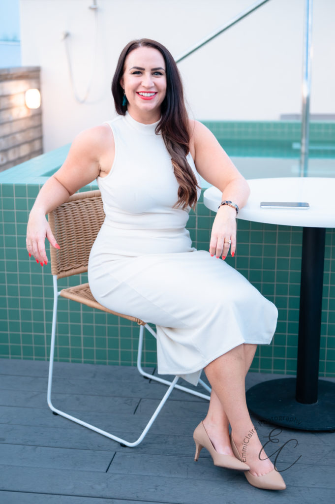 woman in white two-piece outfit sits in a chair in front of rooftop pool while smiling at the camera