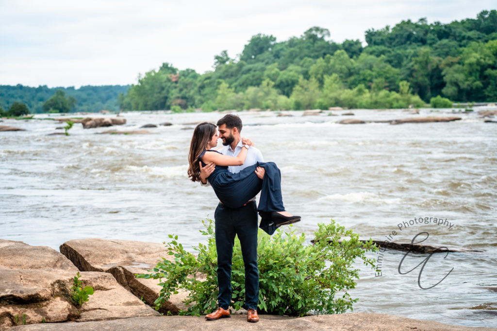 a man in a white shirt and navy blue dress pants holds his fiance in his arms as they touch noses in front of a river